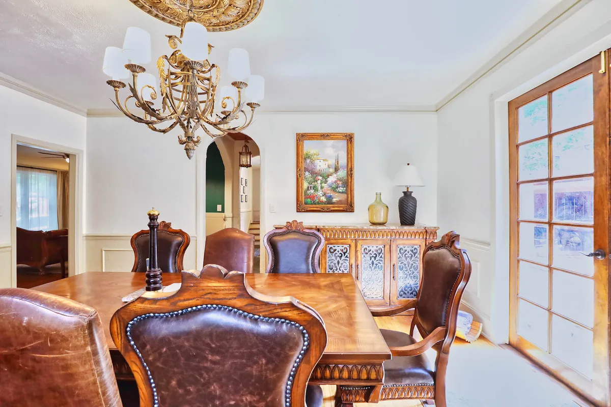 A beautifully appointed dining room featuring a large wooden dining table under an ornate chandelier. The space is accented by a decorative cabinet, elegant wall art, and French doors that allow natural light to flood in, creating a warm and inviting atmosphere ideal for gatherings.