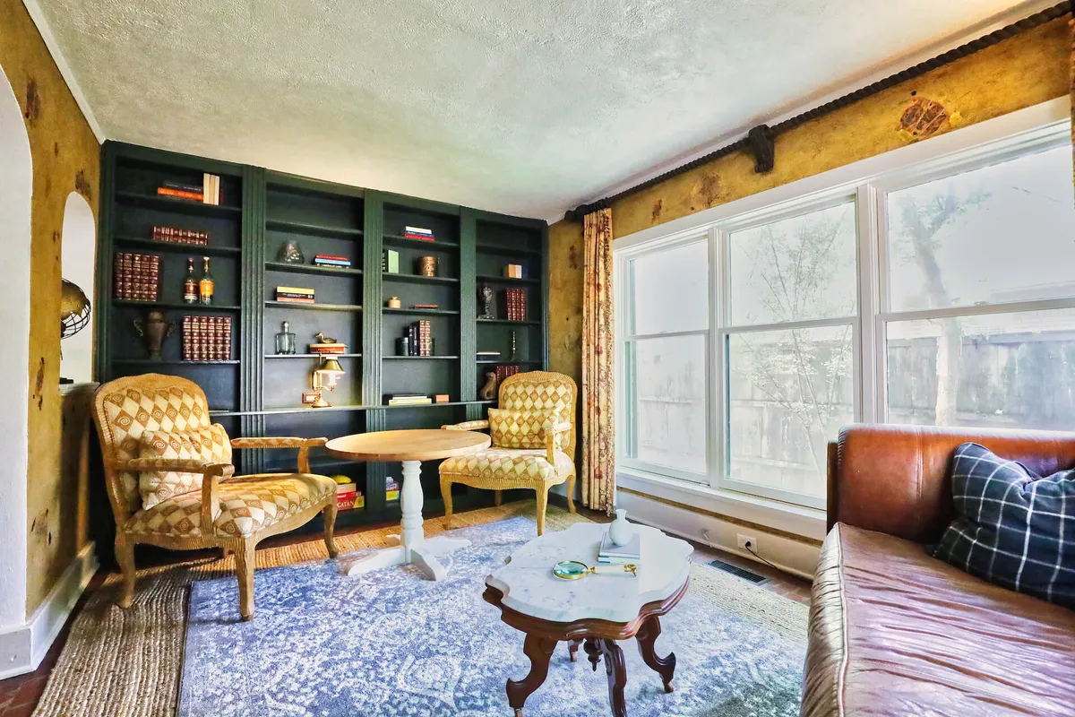 This image features a cozy sitting area with a tall bookshelf filled with books and decorative objects, two patterned chairs arranged for conversation, a small coffee table, and a large window streaming in natural light. The scene feels tranquil and encourages both relaxation and productivity, making it ideal for quiet reading or informal meetings.