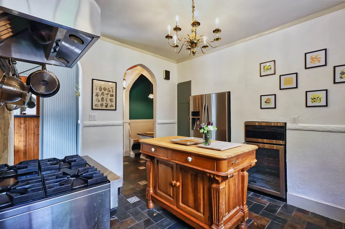 A welcoming kitchen scene featuring a wooden kitchen island, a gas stove, a statement chandelier above, and framed artwork on the wall. The space is arranged for both cooking and entertaining, with ample surfaces for meal prep and gathering.
