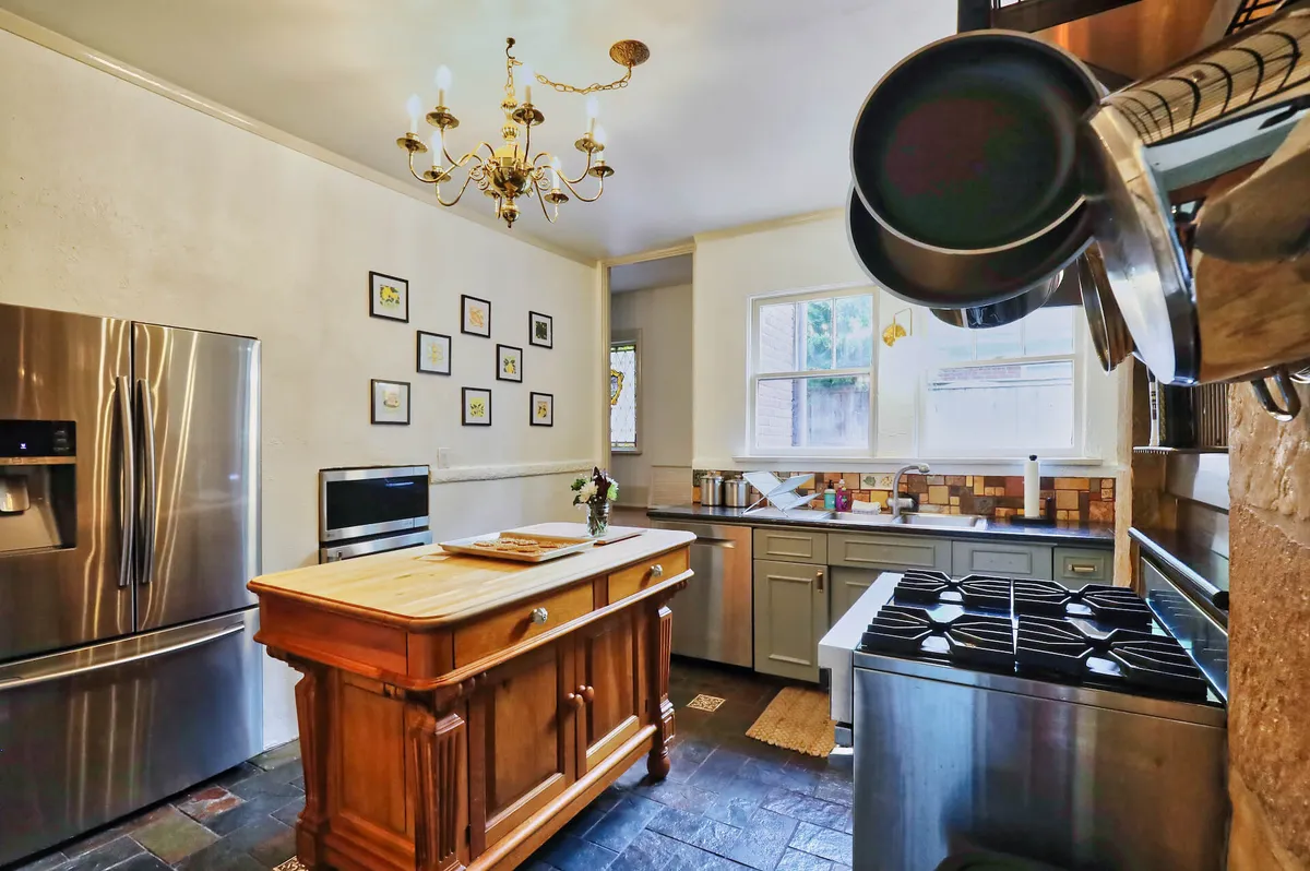 A spacious and inviting kitchen with a central wooden island, modern stainless steel appliances, and a large chandelier overhead. The space is decorated with wall art, offering a warm, communal area ideal for cooking and gathering.
