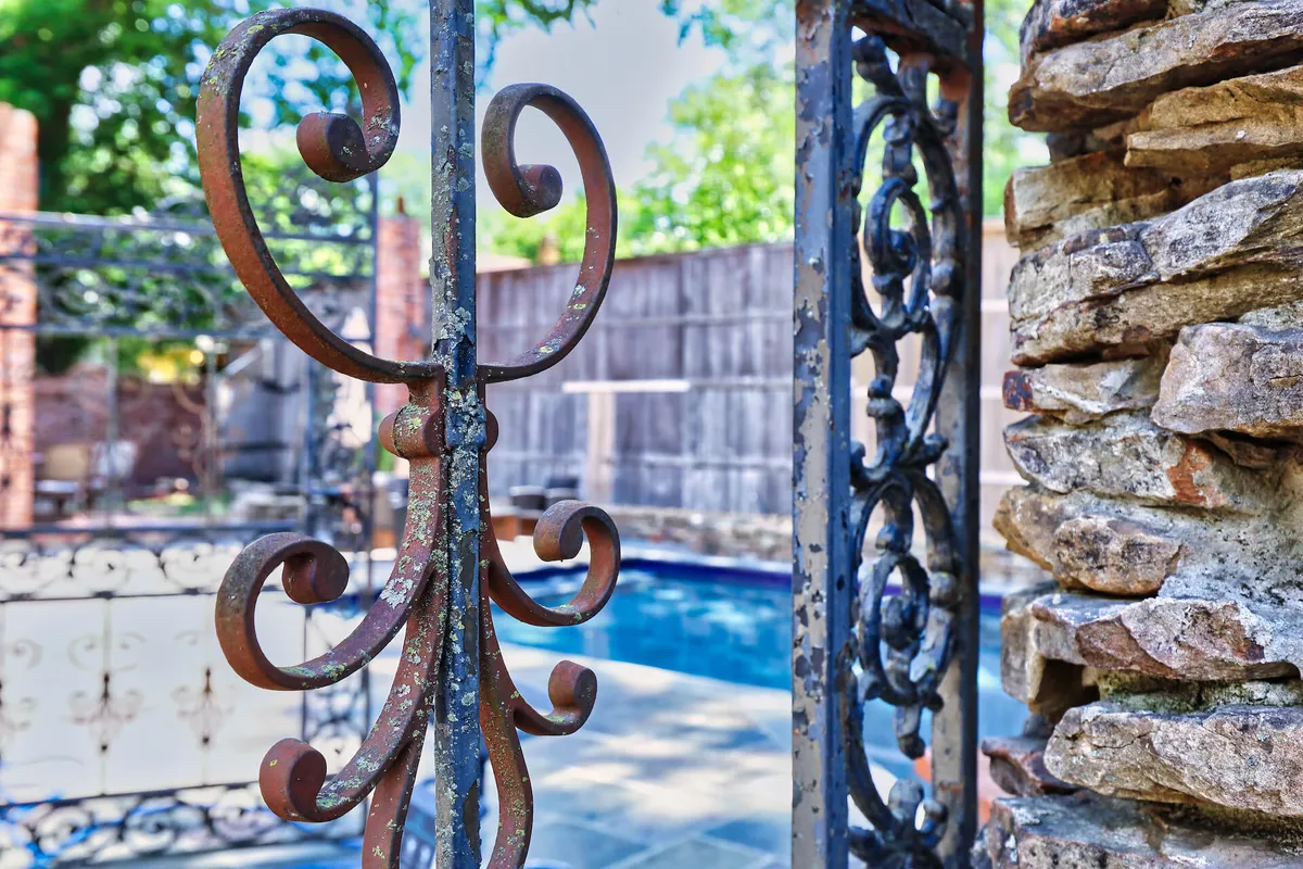 The image showcases a tranquil pool area framed by a decorative wrought-iron gate and stone wall, with the pool glimmering in the background. The mood is serene and secure, ideal for families seeking a peaceful retreat close to city excitement.