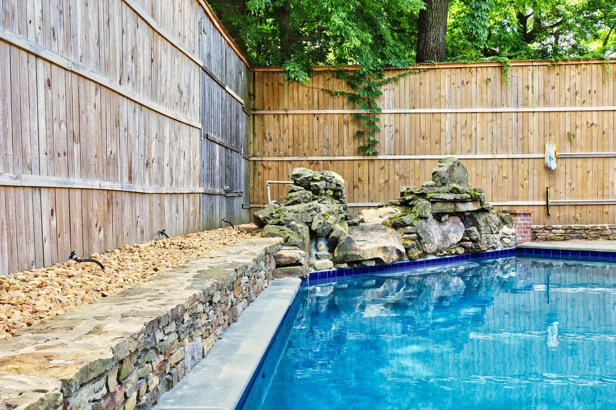A serene pool area featuring a sparkling saltwater swimming pool, bordered by natural rock formations and enclosed by a tall, wooden privacy fence. The setting is tranquil, with opportunities for swimming, sunbathing, or relaxing poolside, and the ambiance hints at both daytime enjoyment and evening swims under the stars.