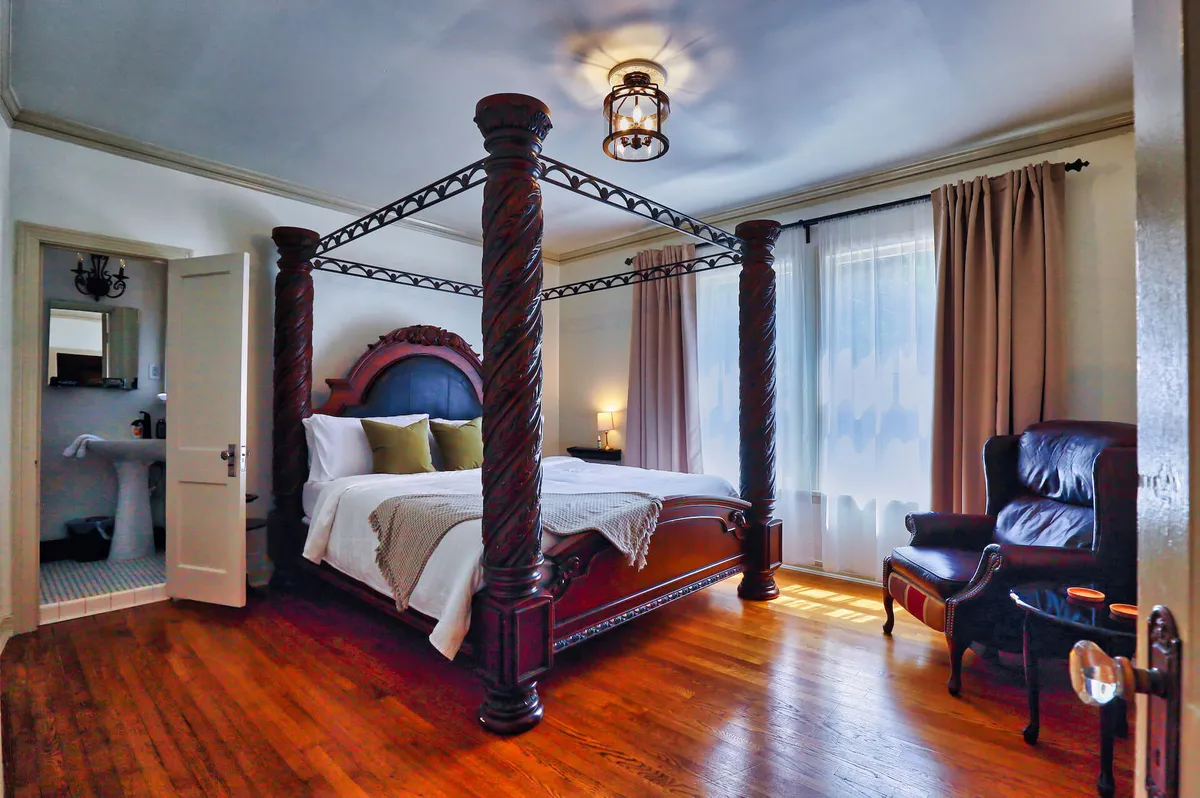 A spacious bedroom featuring a canopy king bed draped in soft linens, set atop wooden flooring. An inviting armchair sits near floor-length curtains, allowing natural light to filter in, creating a cozy and restful atmosphere.