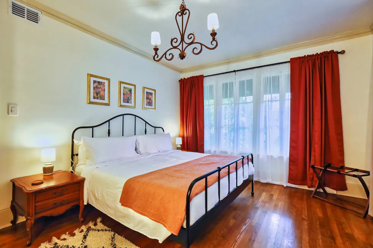 A serene bedroom with a king-sized bed, elegant chandelier, soft curtains, and tasteful artwork. A nightstand sits nearby, perfect for enjoying coffee or conversation in a calm, sunlit setting.