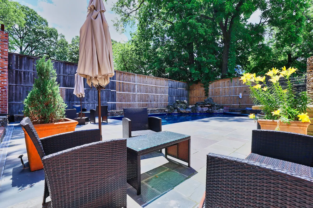 A serene outdoor space with a sparkling swimming pool surrounded by flower pots, a shaded umbrella, and cushioned outdoor seating. The scene evokes relaxation and leisure, suggesting both private retreats and opportunities for social gatherings.