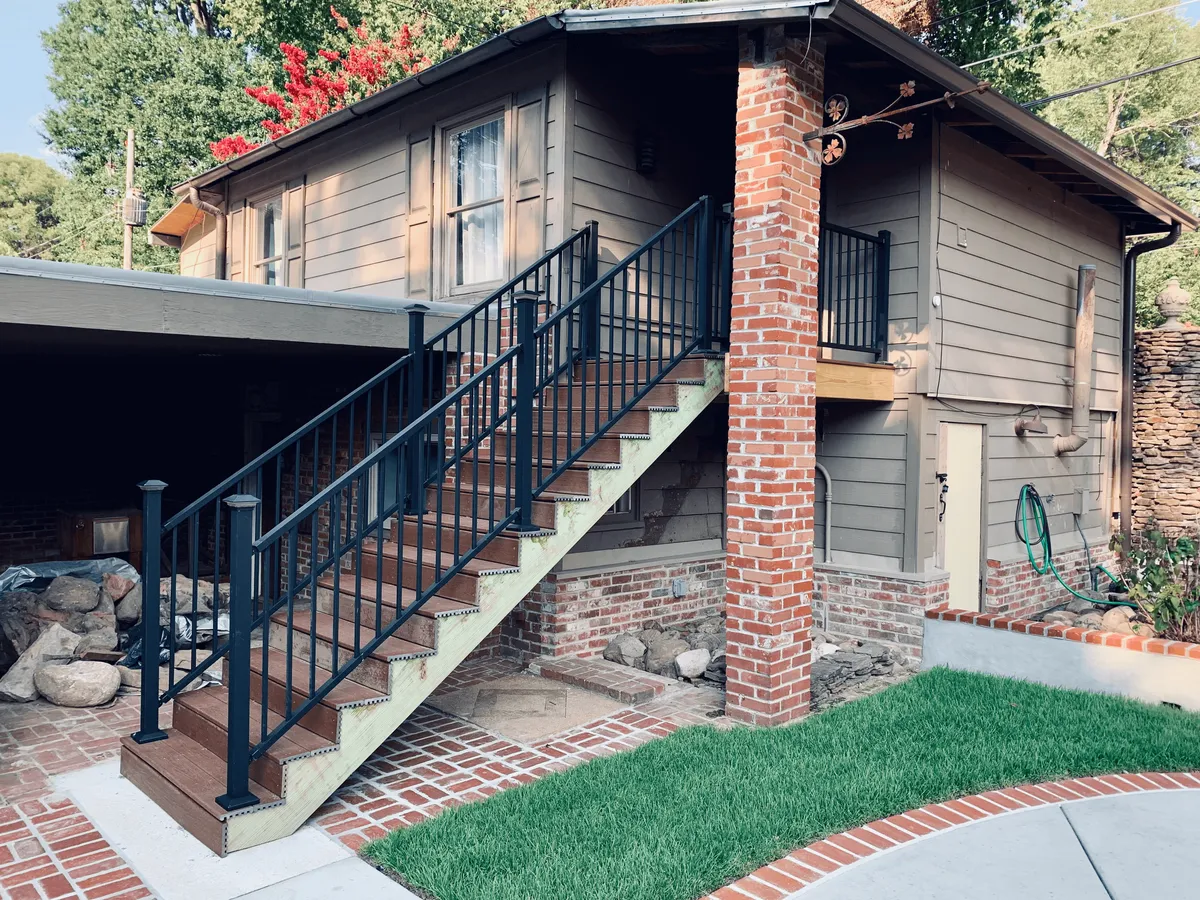A charming exterior view of the historic home featuring a classic brick column, a welcoming staircase, and lush green grass. The window with curtains hints at the cozy interior while the porch and garden area offer a sense of arrival and outdoor living.