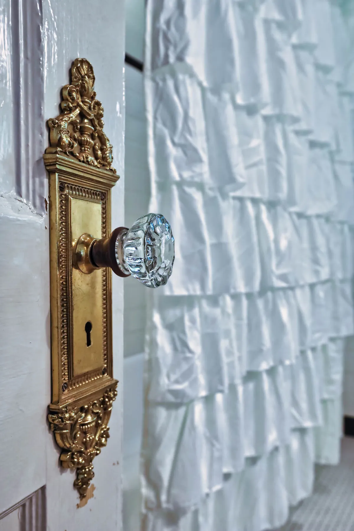 A close-up of an ornate door handle with an intricate door plate, complemented by a white ruffled curtain, suggesting the threshold between public and private space. The image exudes sophistication and anticipation, hinting at the elegance found within.