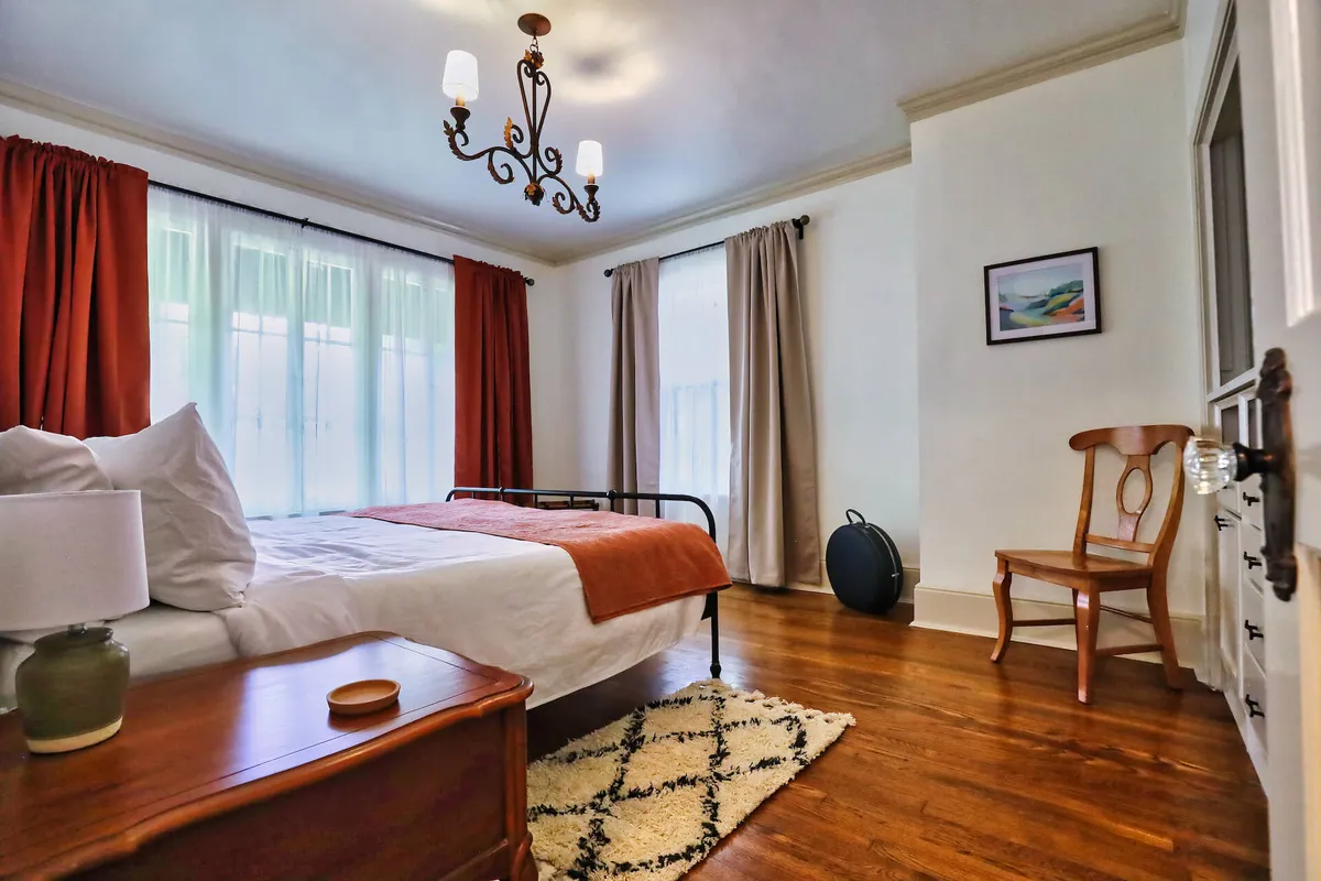 A sunlit bedroom featuring a plush bed adorned with soft linens, elegant chandelier overhead, sheer window curtains letting in natural light, a classic wooden chair, and a richly patterned rug—all creating an inviting, serene atmosphere.