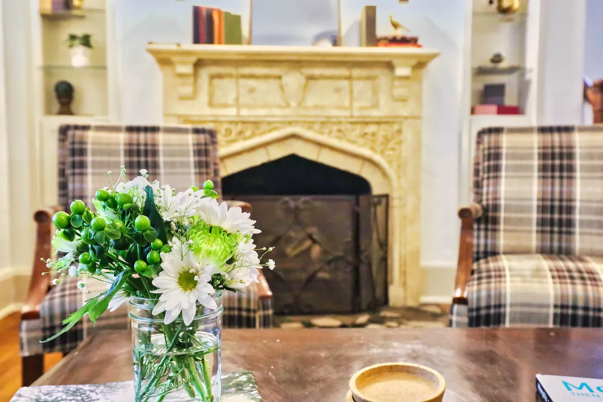 A cozy living room scene centered around a fireplace, with plaid armchairs and a vibrant floral arrangement adding comfort and color. The setting encourages relaxation, conversation, and togetherness, with the warmth of the fire emphasizing the room’s inviting nature.
