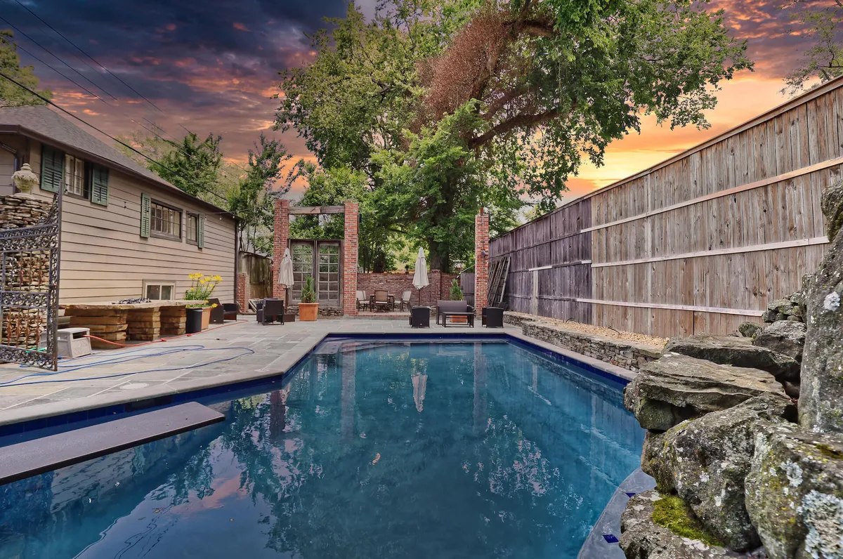 A tranquil exterior view of the pool area, complete with lush greenery, a decorative stone wall, and a cozy outdoor seating zone. The mood is serene and relaxed, perfect for unwinding or entertaining, with the pool as the central feature.
