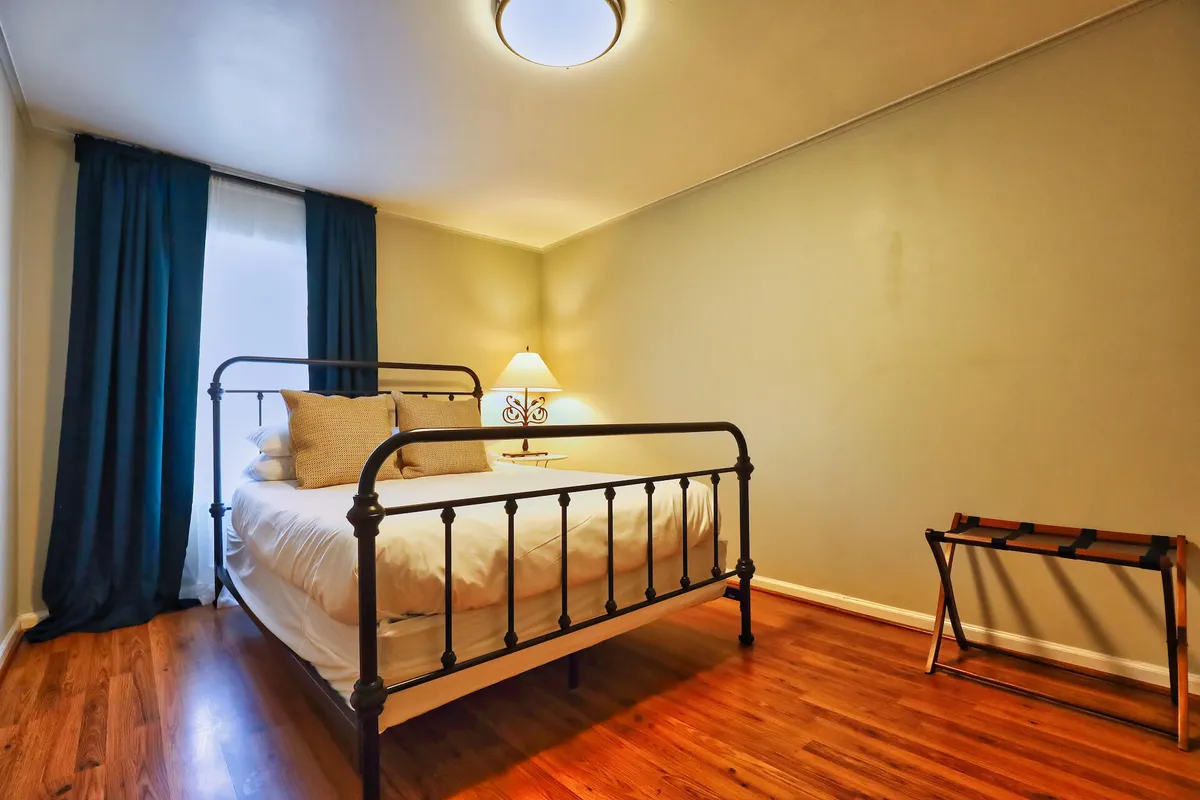 A serene bedroom scene with a neatly made bed, soft lamp lighting, curtains gently diffusing sunlight, and warm wooden flooring. The ambiance is peaceful and inviting, ideal for restful nights or quiet moments of reflection.