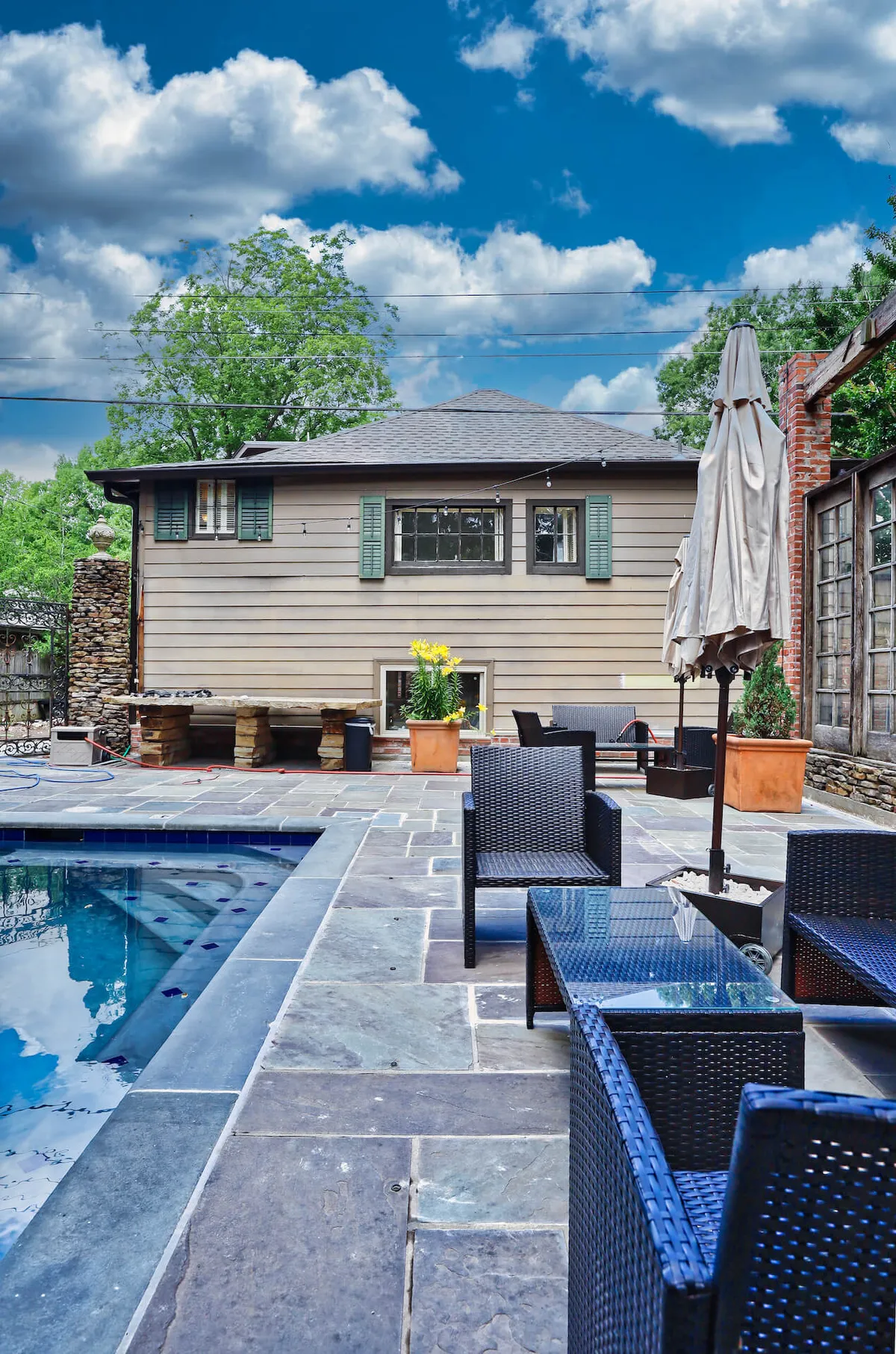 A stylish poolside scene with a spacious swimming pool, inviting outdoor seating area, and the house’s charming facade in the background. Lush planters and flowers add to the resort-like atmosphere, while the arrangement suggests both daytime lounging and socializing during sunset hours.
