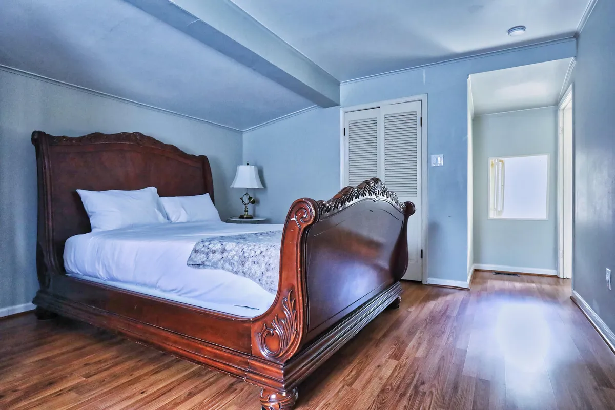 An ornate wooden bed with crisp white linens and a classic nightstand with a lamp, set against a backdrop of soft light. The calm scene is inviting for rest, reading, or simply enjoying a peaceful moment.