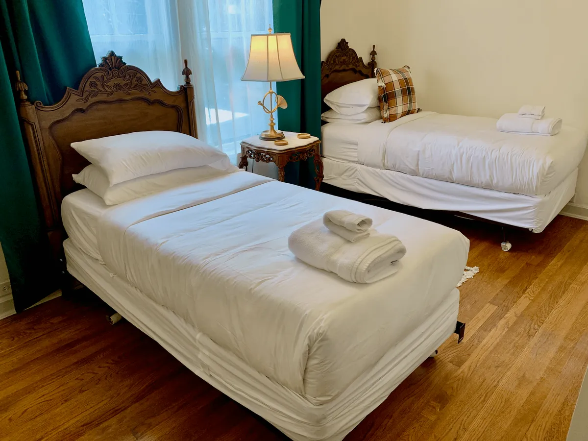 A warm and homey bedroom with wooden bed frames, plaid accent pillow, soft bedside lamp, and neatly arranged towels. Sunlight streams in, highlighting the inviting bedding and encouraging relaxation or conversation.